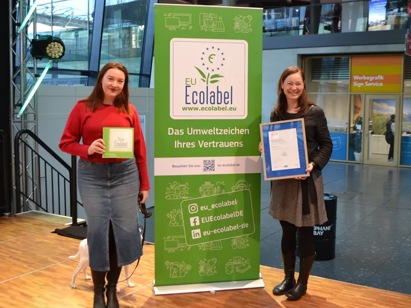 Kristina Kuhn, Inhaberin des Kerlin, links im roten Pullover mit der EU-Ecolabel-Plakette und einem weißen Hund an der Leine. Rechts Dr. Kristin Stechemesser vom Umweltbundesamt mit der gerahmten Urkunde, dazwischen ein Aufsteller des EU Ecolabels.