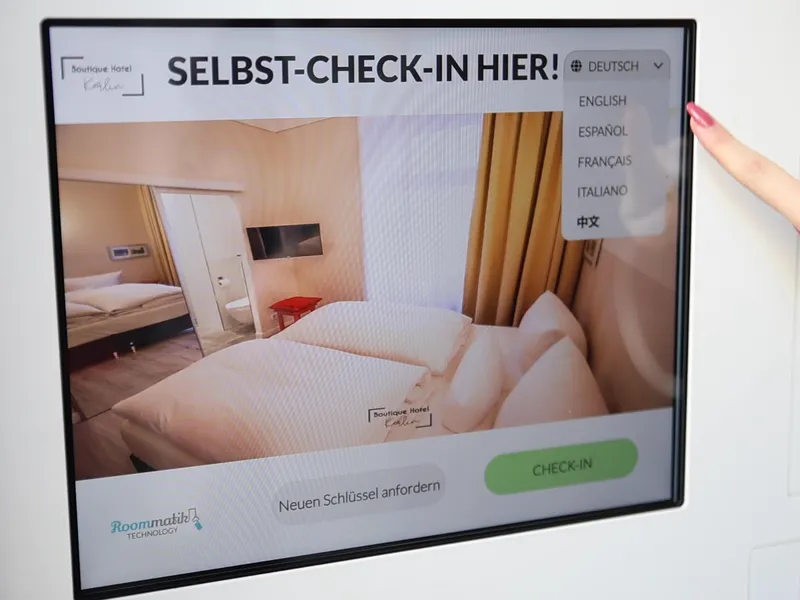 Der Self-Check-in-Kiosk des Kerlin: Startbildschirm „Selbst-Check-in hier!“ mit geöffneter Sprachauswahl Deutsch, Englisch, Spanisch, Französisch, Italienisch und Chinesisch. Daneben Kartenterminal und Ausgabe für die Schlüsselkarte.