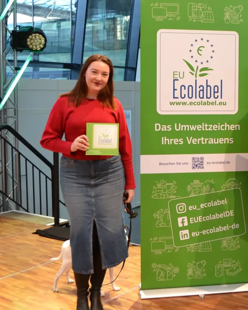 Kristina Kuhn, Inhaberin des Kerlin, im roten Pullover mit der Plakette des EU Ecolabels, daneben ein Aufsteller des EU Ecolabels